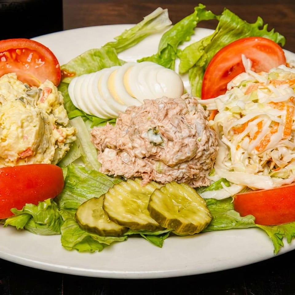 A salad platter with scoops of potato salad, tuna salad, coleslaw, sliced tomatoes, hard-boiled eggs, and pickles.
