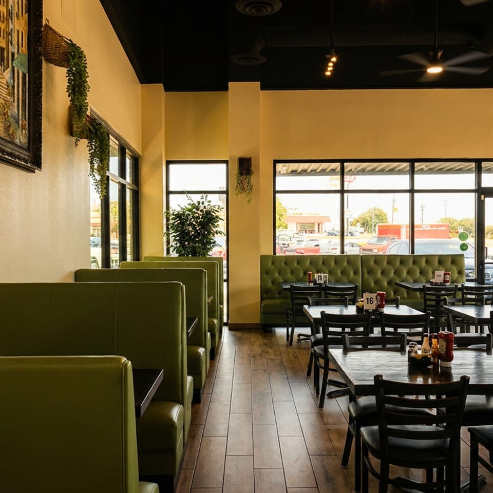 Empty restaurant dining room with green booths, dark tables, and large windows overlooking a parking lot.
